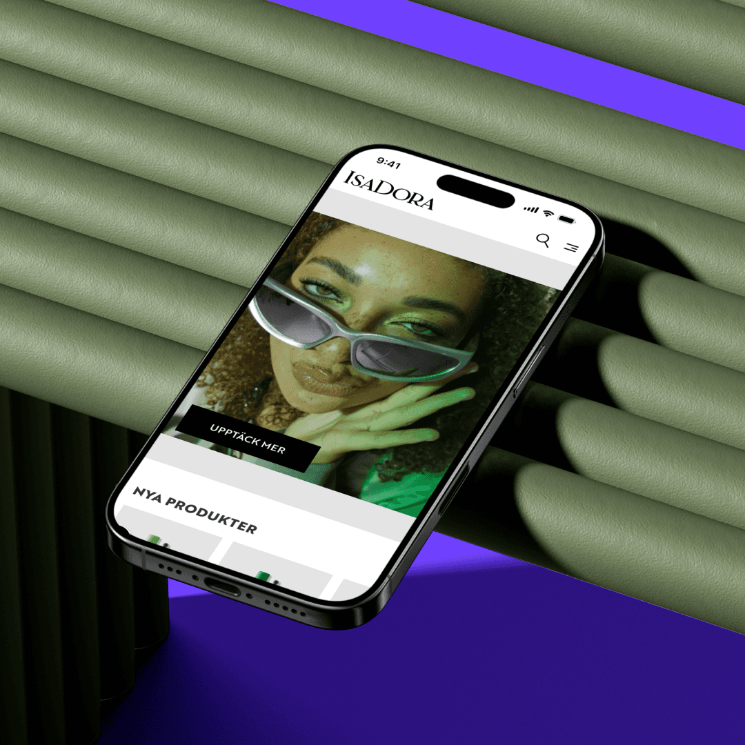 IsaDora Virtual makeup A smartphone displaying IsaDora's website featuring a woman wearing sunglasses. The phone is positioned diagonally on a ribbed green surface with a purple background.